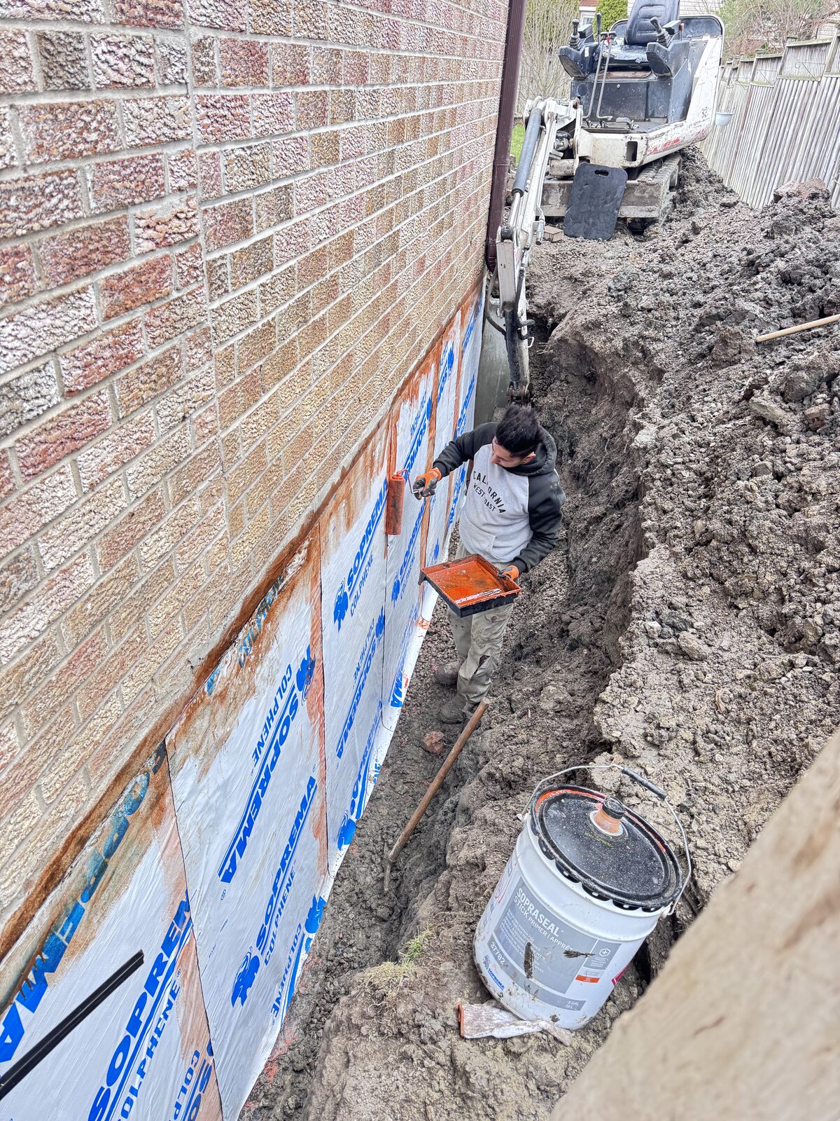 Worker applying waterproofing to a foundation wall with a roller.